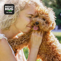 A smiling woman with curly blonde hair hugs a small, fluffy dog close to her face, both appearing happy. The dog, likely a poodle mix, sticks out its tongue playfully. The background features greenery, indicating a sunny outdoor environment. The image also includes the text "Nature's HUG PET FOOD" in the top left corner, suggesting a promotional context.
