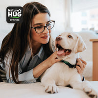 A young woman with long hair and glasses gently interacts with a cheerful, light-colored puppy on a bed. The woman looks at the puppy with affection, while the puppy has its mouth open, appearing to playfully bark. In the corner is a logo for "Nature's Hug Pet Food."