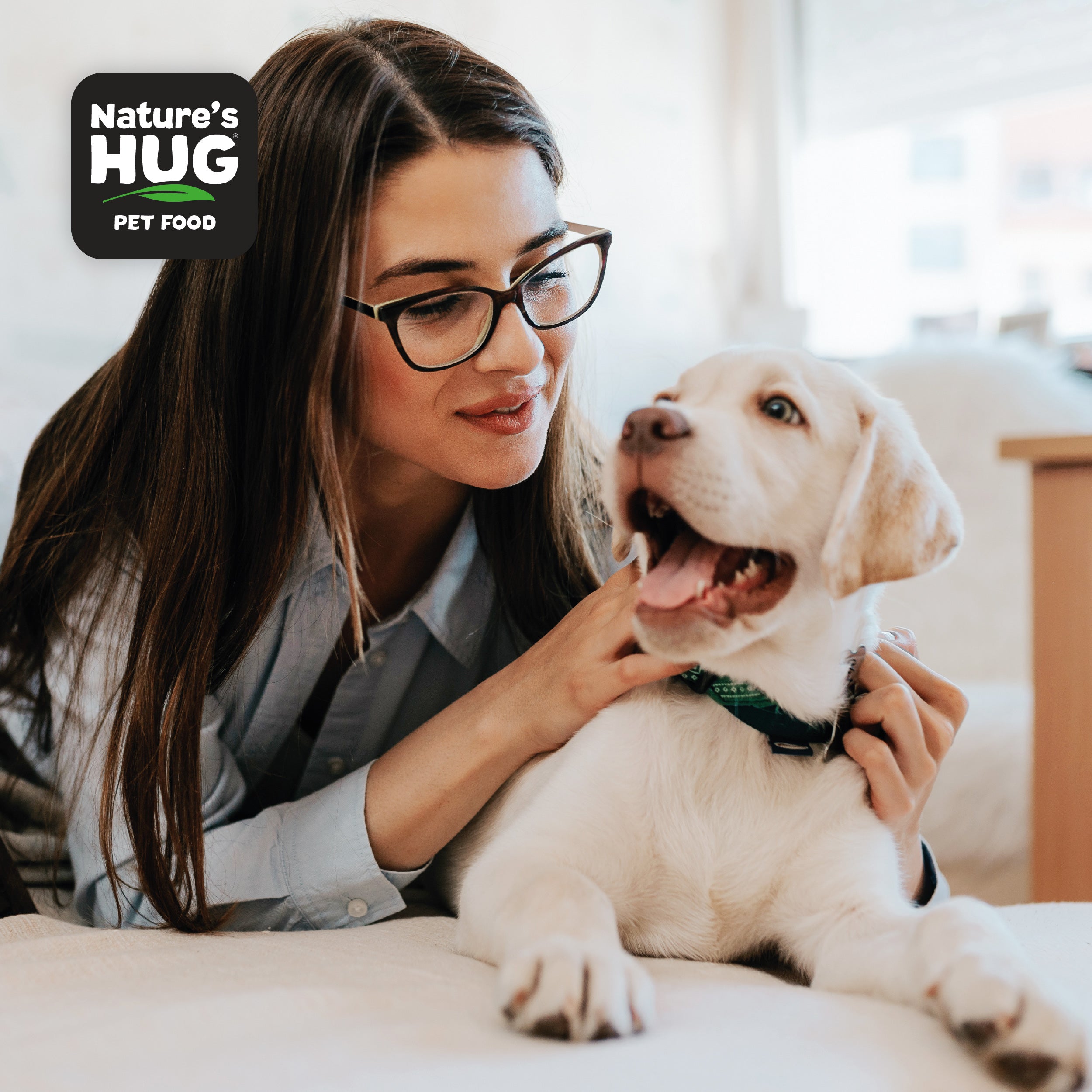 A young woman with long hair and glasses gently interacts with a cheerful, light-colored puppy on a bed. The woman looks at the puppy with affection, while the puppy has its mouth open, appearing to playfully bark. In the corner is a logo for "Nature's Hug Pet Food."