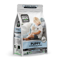 The image features a bag of Puppy, Nutrients for Growth Formula Dog Food for Toy & Small Breeds "Nature's Hug". The packaging highlights that it offers complete and balanced nutrition for growth in the first year. In the foreground, a child with short blonde hair sits on a couch, gently holding a small white puppy. The background shows a cozy setting with patterned cushions.
