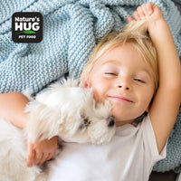A joyful boy lies on a soft, textured blanket, smiling at the camera while hugging a small white dog closely. The boy has light blonde hair and is wearing a white t-shirt. The background features a light blue knitted blanket, enhancing the warm atmosphere of the moment.