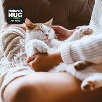 A cozy indoor scene features a person gently holding a sleeping cat in their arms. The cat looks content with its eyes closed. The person is wearing a soft, knitted sweater, and the background includes a plush couch. Visible in the corner is a logo for Nature's Hug Pet Food.