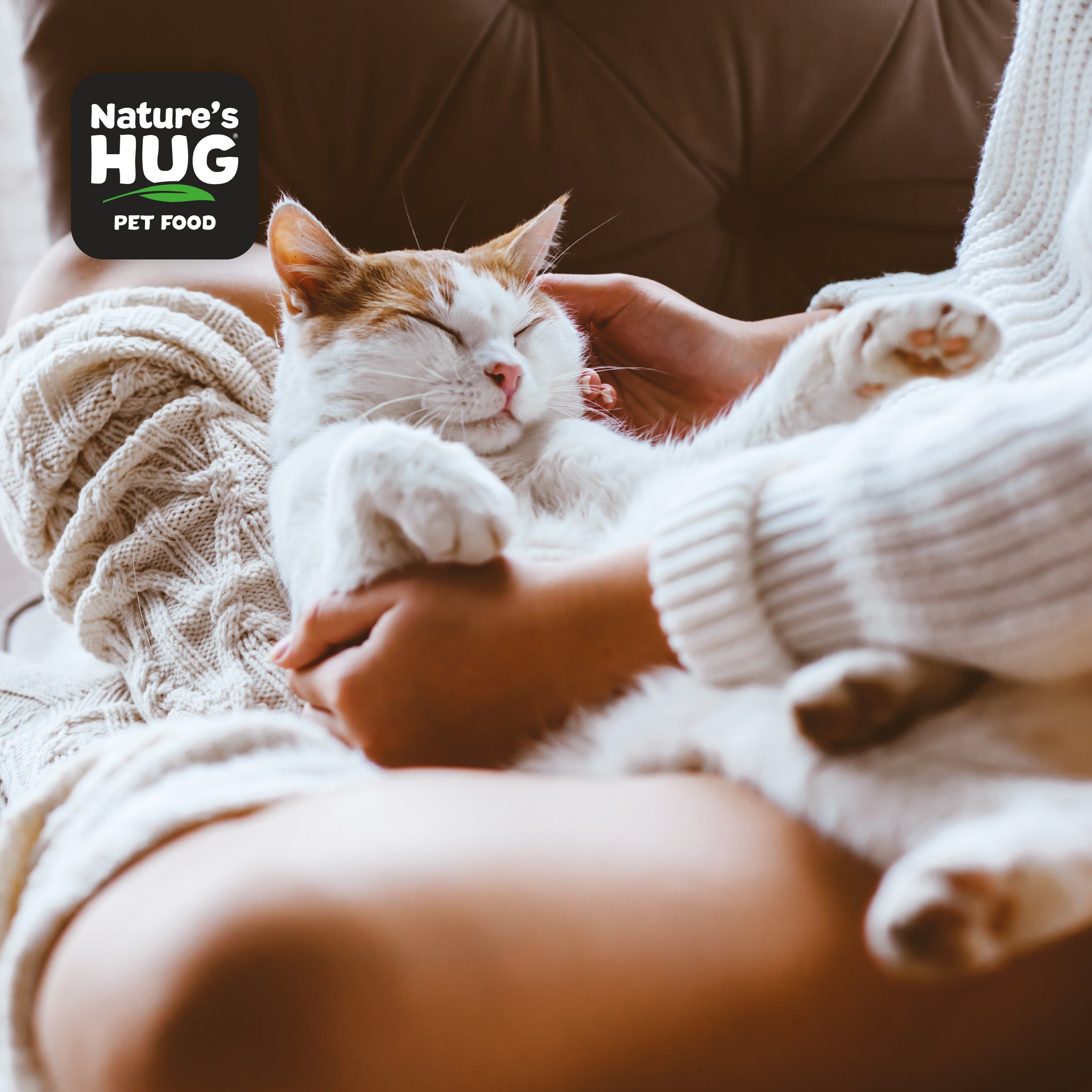 A cozy indoor scene features a person gently holding a sleeping cat in their arms. The cat looks content with its eyes closed. The person is wearing a soft, knitted sweater, and the background includes a plush couch. Visible in the corner is a logo for Nature's Hug Pet Food.