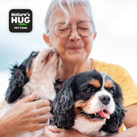 An older woman with glasses smiles gently while holding two dogs in her arms. One dog is licking her cheek, showcasing affection, while the other sits contentedly beside her. The image includes the "Nature's Hug" pet food logo in the top left corner.