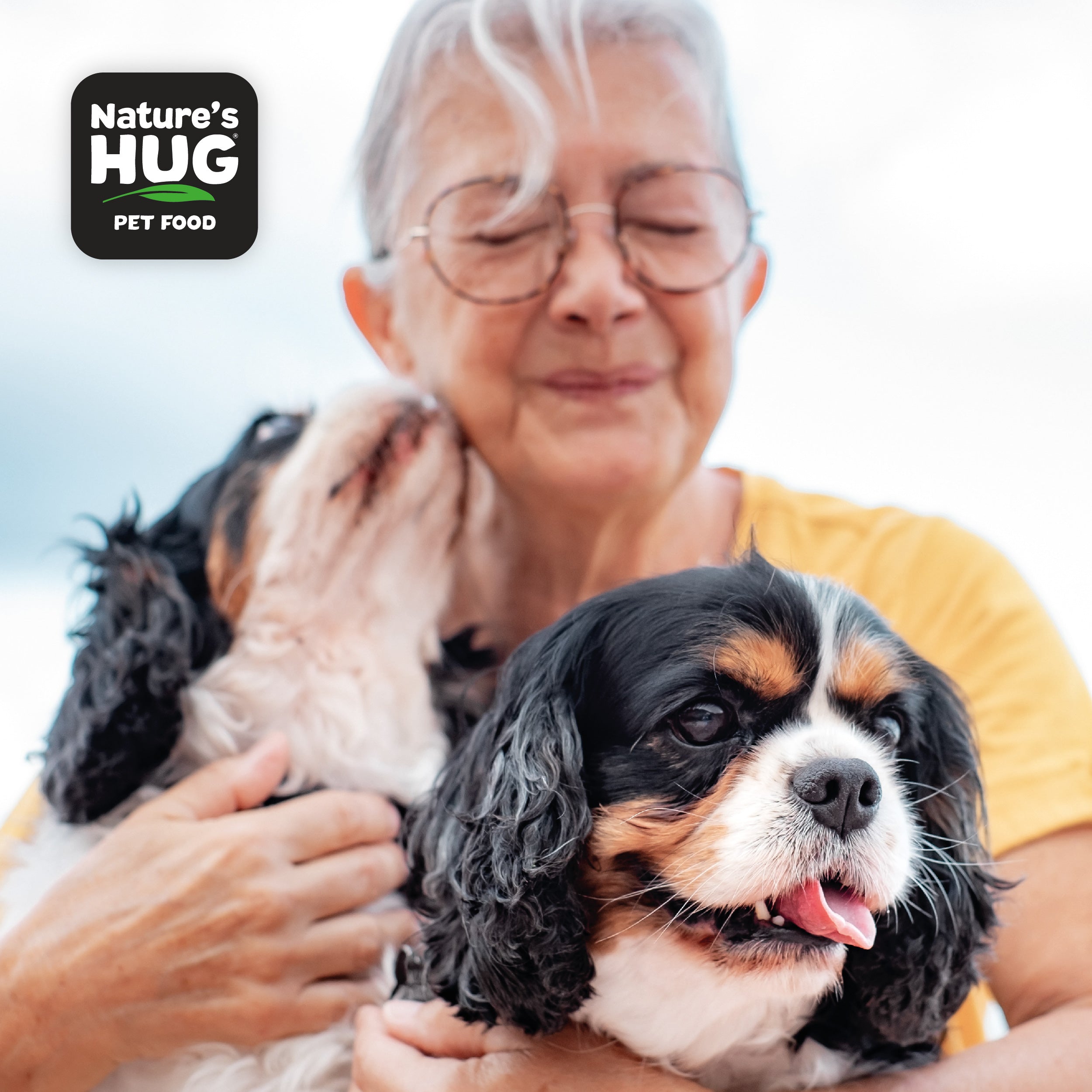 An older woman with glasses smiles gently while holding two dogs in her arms. One dog is licking her cheek, showcasing affection, while the other sits contentedly beside her. The image includes the "Nature's Hug" pet food logo in the top left corner.
